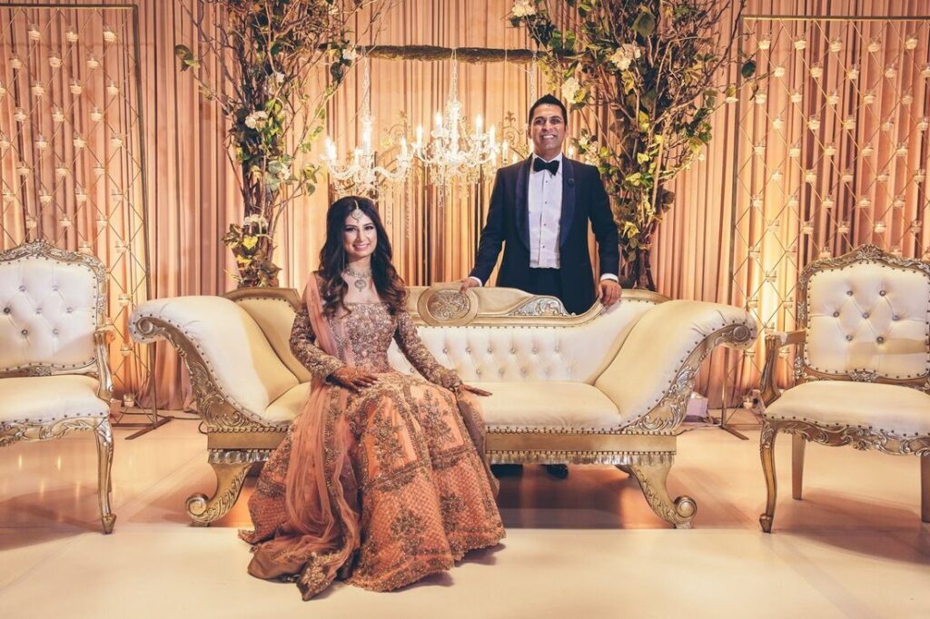 Indian wedding reception scene featuring a groom in a tailored black-tie ensemble beside the bride, illustrating elegant men’s reception outfits such as classic tuxedos and refined spezzato styling for formal wedding celebrations.